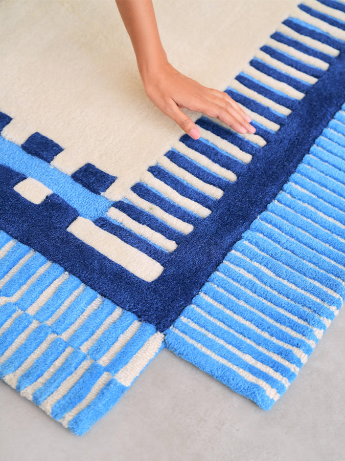 Hand touching Nested rug's raised ivory ladder pattern, navy border and sky blue striped corner